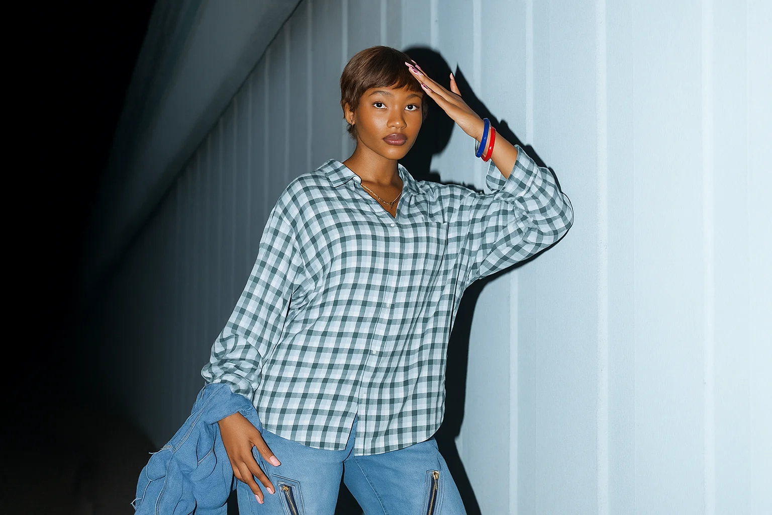 Foto de moda de uma mulher com camisa xadrez e calça jeans posando à noite em frente à parede